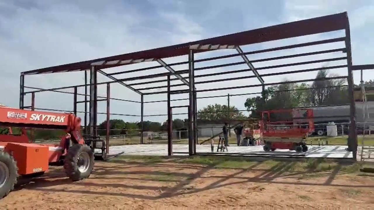 A red iron rigid frame standing on its slab, columns and rafters bolted up, before any sheeting